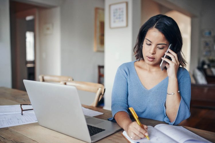 Woman sitting at table on the phone taking notes beside an open laptop