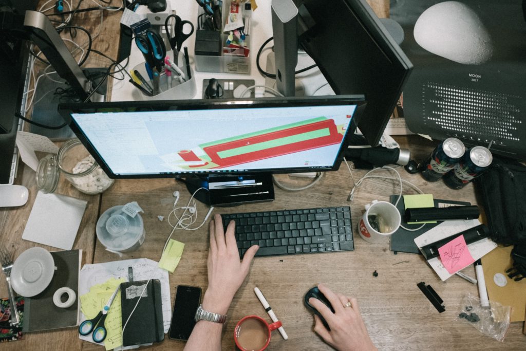 Aerial view of a cluttered workspace with a person's hands on their keyboard and mouse