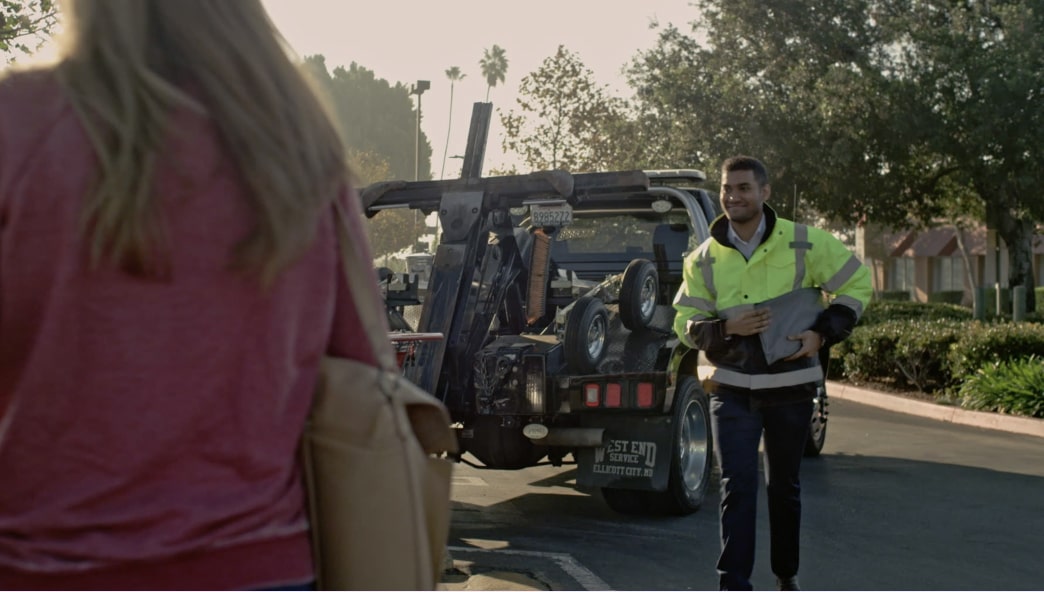 Worker holding clipboard approaching woman with tow truck in background