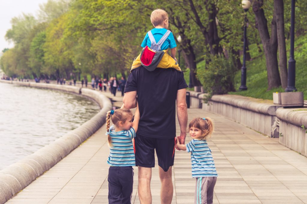 Father with three children walking in a park (one on shoulders, and two on either side holding his hands)